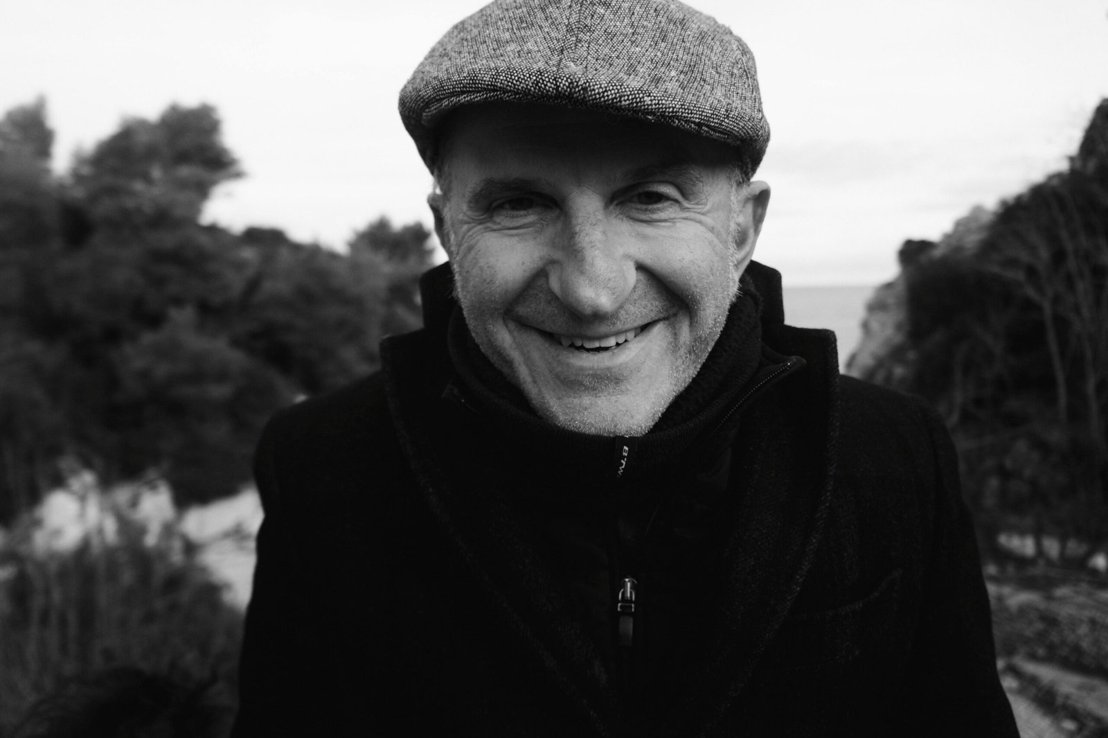 Black and white portrait of a smiling senior man wearing a flat cap outdoors.